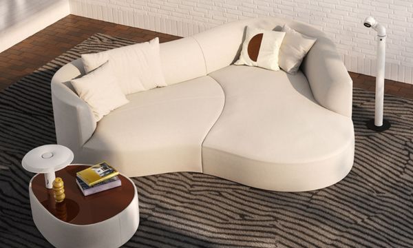 Ein modernes, geschwungenes weißes Sofa mit beigen und gemusterten Kissen steht auf einem gestreiften Teppich neben einem kleinen Couchtisch mit Büchern und einer Lampe vor einer weißen Backsteinwand - die perfekte Inspiration vom Möbelhaus Bad Homburg.