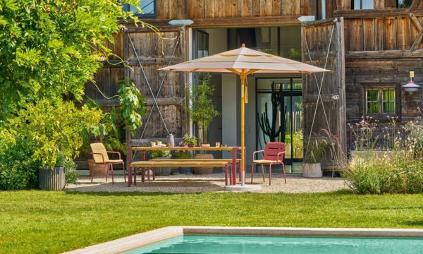 mdm_juli_26_weishaeupl_teakschirm-1 In einem idyllischen Garten steht der Teakholz-Sonnenschirm von Weishäupl an einem Pool. Im Hintergrund ist eine Holzhütte zu sehen.