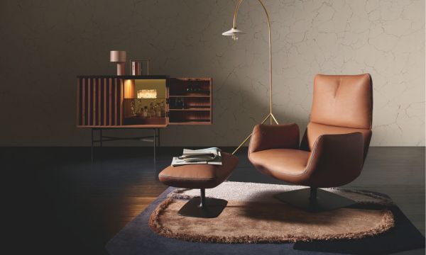 Ein modernes Wohnzimmer mit einem braunen Ledersessel, einer passenden Ottomane, einem Sideboard mit Ambientebeleuchtung, einer Stehlampe und einem runden Teppich auf dunklem Boden - perfekt gestylt wie ein Showroom im Möbelhaus Bad Homburg.