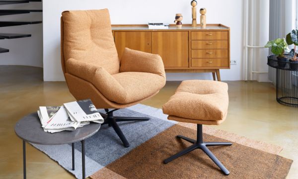 In einem modernen Wohnzimmer stehen ein orangefarbener Polstersessel mit passender Ottomane vom Möbelhaus Bad Homburg, ein Sideboard aus Holz, ein Couchtisch mit Zeitschriften und ein gemusterter Teppich. In einem modernen Wohnzimmer stehen ein orangefarbener Polstersessel mit passender Ottomane vom Möbelhaus Bad Homburg, ein Sideboard aus Holz, ein Couchtisch mit Zeitschriften und ein gemusterter Teppich.