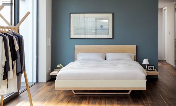 Minimalistisches Schlafzimmer mit einem Holzbett vom Möbelhaus Bad Homburg, weißem Bettzeug, einer grauen Akzentwand, gerahmten Kunstwerken und einem Kleiderständer an der Seite. Durch große Fenster fällt natürliches Licht ein.