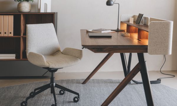 Ein moderner Holzschreibtisch mit einem beige gepolsterten Drehstuhl, einem Regal, Büchern, einer Lampe und einem Teppich in einer minimalistischen Home-Office-Einrichtung vom Möbelhaus Bad Homburg.