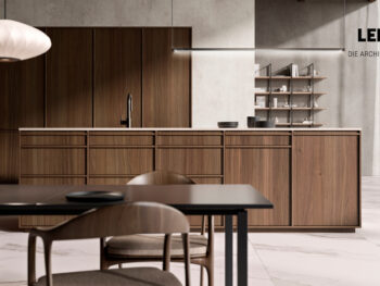 Moderne Küche mit Holzschränken, Kochinsel und minimalistischem Einrichtungsstil. Im Vordergrund stehen ein schwarzer Esstisch und Stühle. Sonnenlicht fällt durch transparente Vorhänge. Zu sehen ist ein Design von Original Braum Interior Design, LEICHT. An der Wand hängt „DIE ARCHITEKTURKÜCHE“.