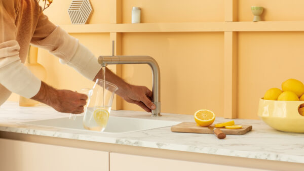 Jemand füllt einen Glaskrug mit Wasser aus einem Quooker-Wasserhahn; darin schwimmen Zitronenscheiben, auf der Arbeitsplatte liegen frische Zitronen und ein Schneidebrett.