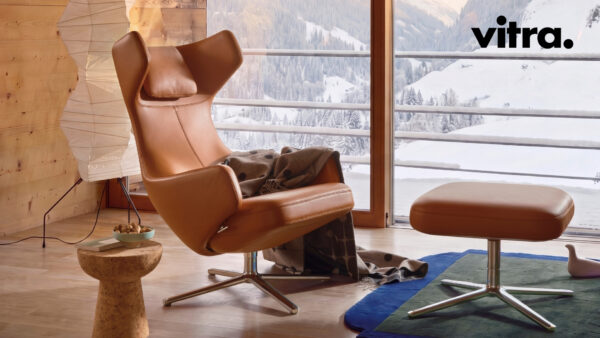 Ein brauner Ledersessel mit passendem Hocker steht vor einem Fenster mit Blick auf verschneite Berge; über den Sessel ist eine Decke drapiert. Inspiriert von „gutes Design“ und vorgestellt in den „Vitra Home Stories“, Winter 2026/2027, strahlt die Szene Wärme und zeitlosen Stil aus. Das Vitra-Logo befindet sich in der oberen rechten Ecke.