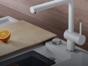 Eine moderne Blanco Linus Armatur in Weiß steht neben einem Waschbecken, ergänzt durch ein hölzernes Schneidebrett mit einer halbierten Orange.