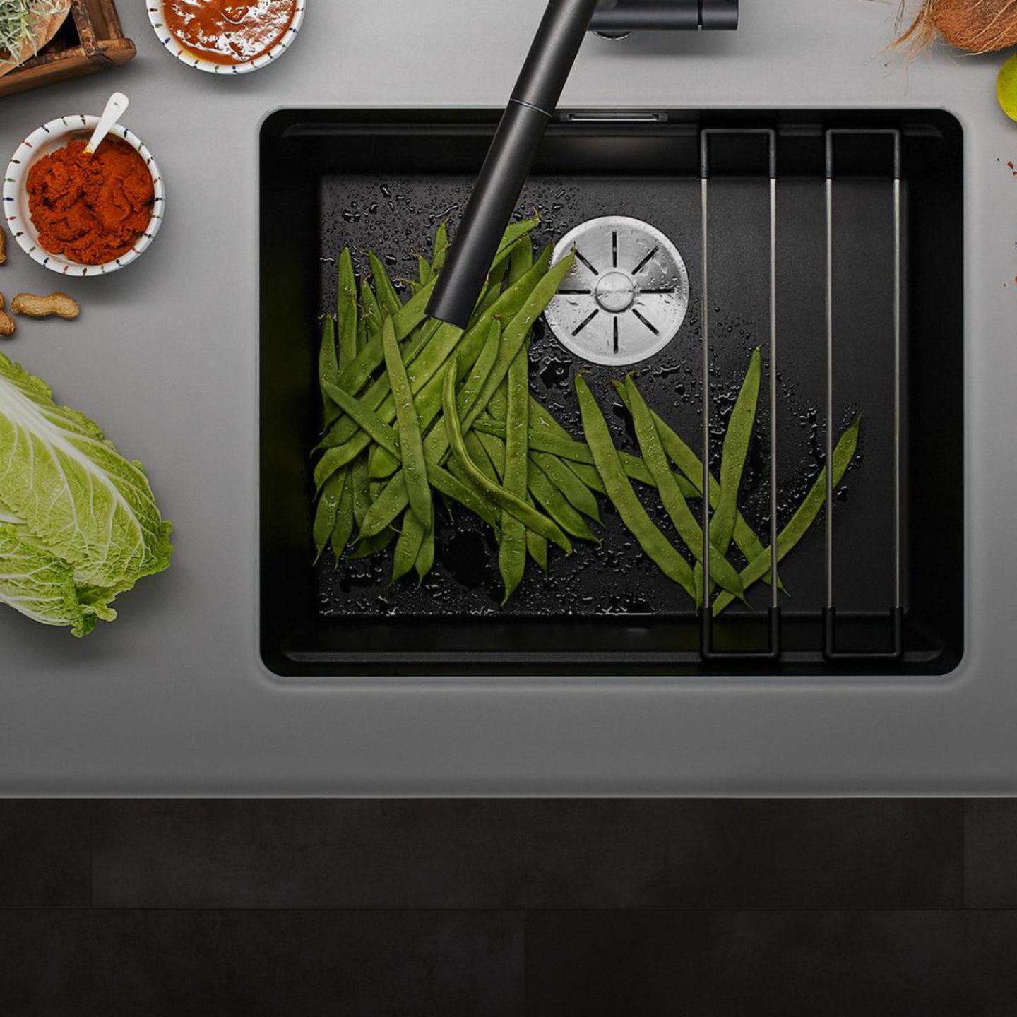 Blick von oben auf grüne Bohnen, die in einer schwarzen Blanco Etagon Spüle gewaschen werden, umgeben von Gemüse und kleinen Schalen mit Gewürzen auf einer grauen Arbeitsplatte.