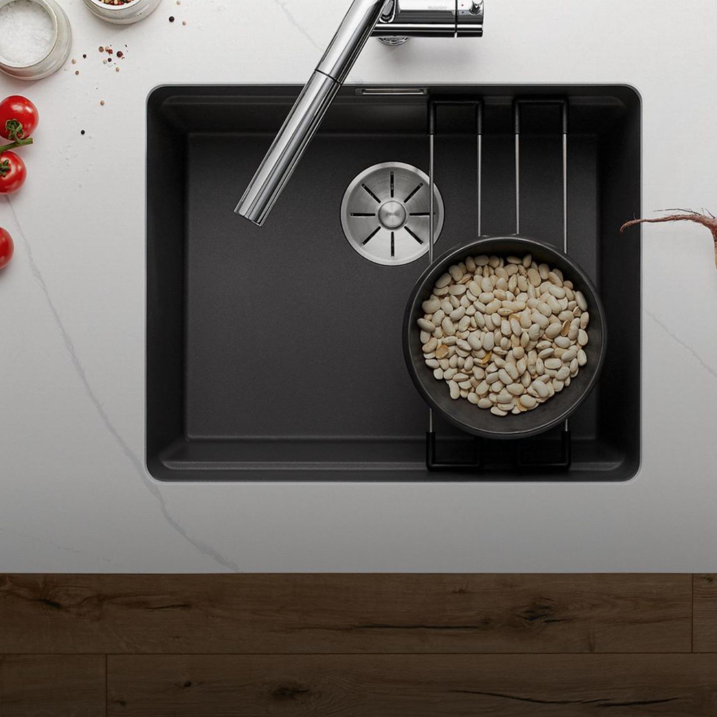 Eine Blanco Etagon Spüle mit einem Wasserhahn aus rostfreiem Stahl, mit einem Topf weißer Bohnen auf einem Gestell über der Spüle, neben Tomaten, Gewürzen und Kräutern, die fein säuberlich auf der weißen Arbeitsplatte angeordnet sind.