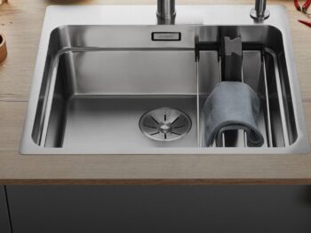Eine moderne Blanco Etagon Spüle aus Edelstahl mit einem Wasserhahn, einem Abfluss, einer Trennwand und einem über einen Halter drapierten Tuch. Lebensmittel und Utensilien sind auf der umliegenden Arbeitsplatte sichtbar.