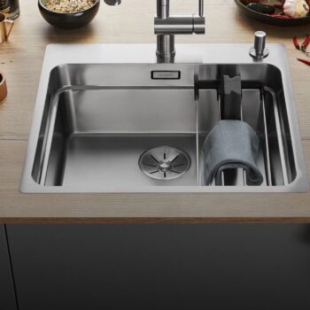 Eine moderne Blanco Etagon Spüle aus Edelstahl mit einem Wasserhahn, einem Abfluss, einer Trennwand und einem über einen Halter drapierten Tuch. Lebensmittel und Utensilien sind auf der umliegenden Arbeitsplatte sichtbar.
