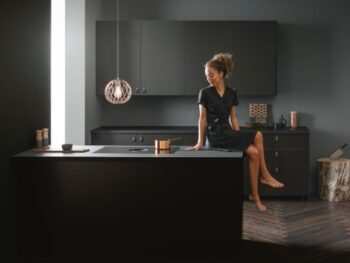 Eine Frau in einem schwarzen Kleid sitzt an einer modernen schwarzen Kücheninsel in einer minimalistischen, dunkel getönten Küche mit Holzboden und moderner Beleuchtung.