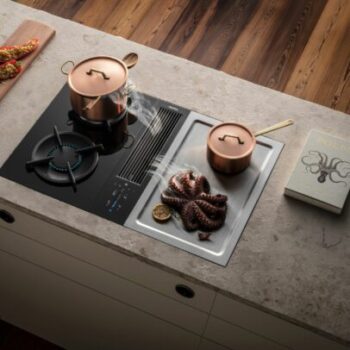 Eine moderne Küchenarbeitsplatte mit einem Bora Classic 2.0 Gasbrenner, Kochtöpfen und gegrilltem Oktopus auf einem Metalltablett neben einem Kochbuch.