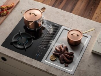 Ein modernes Bora-Kochfeld mit einem Topf auf einem Gasbrenner, ein weiterer Topf, ein Oktopus, der auf einer Grillplatte gegrillt wird, und ein Kochbuch mit dem Titel Polpo auf dem Tresen.