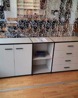 Ein Dauphin Home Sideboard mit zwei Schränken, vier Schubladen und offenen Regalen steht auf einem Holzfußboden, ergänzt durch einen dekorativen weißen Hängetrenner im Hintergrund.
