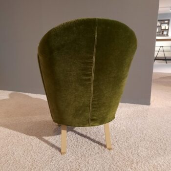 Ein grüner Brühl-Samt-Sessel mit goldenen Beinen steht auf einem hellen Teppich, von hinten betrachtet, in einem modernen Innenraum.