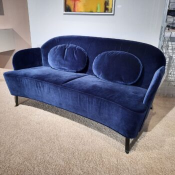 Ein modernes blaues Samtsofa mit zwei großen ovalen Rückenkissen auf schwarzen Beinen, platziert auf einem beigen Teppich in einem zeitgenössischen Interieur.