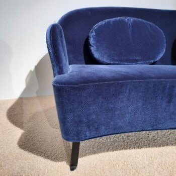 Nahaufnahme eines blauen Samtsofas mit abgerundeten Kanten und einem passenden ovalen Kissen, das auf einem beigen Teppich steht.