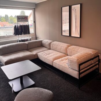 Ein modernes Wohnzimmer mit einem beigen Brühl-Ecksofa, einem schwarzen Couchtisch, gerahmten abstrakten Kunstwerken an der Wand und einer Stoffmusterauslage neben einem großen Fenster – schlichte Möbelstücke, die sorgfältig ausgewählt wurden, um einen zeitlosen Stil zu schaffen, der ewig Bestand hat.