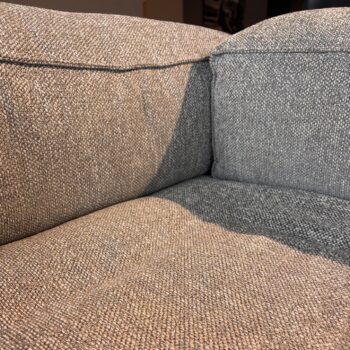 Nahaufnahme der Ecke eines Sofas aus grauem, strukturiertem Stoff, wie beispielsweise der „Brühl Ecksofa Easy“-Modelle, die die Sitz- und Rückenkissen mit sichtbaren Nähten und Schatten zeigt – ein einladender Anblick, der den Eindruck vermittelt, als könnte er ewig so bleiben.
