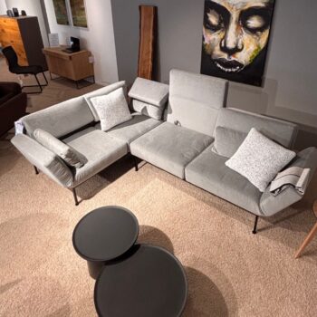 Ein graues Brühl-Roro-Modulsofa mit vier weißen Kissen, zwei runden schwarzen Couchtischen, einem beigen Teppich und einem abstrakten Gesichtsbild an der Wand in einem modernen Wohnzimmer.