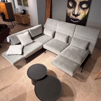 In einem Wohnzimmer steht ein modernes graues Brühl-Sofamodul mit zwei gemusterten Kissen neben zwei runden schwarzen Couchtischen auf einem beigen Teppich. An der Wand darüber hängt ein großes Gemälde, das das elegante, sanfte Design des Raums unterstreicht.