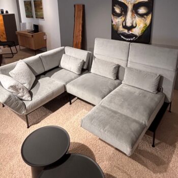 Ein hellgraues Brühl-Ro-Ro-Ecksofa mit zahlreichen Kissen ist in L-Form auf einem beigen Teppich angeordnet, davor stehen zwei runde schwarze Tische und an der Wand dahinter hängt ein großes Gemälde.