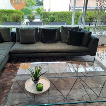 Ein modernes Wohnzimmer mit einem Charles Carré Sofa von B&B Italia, einem Couchtisch aus Glas, einer kleinen Pflanze, einer Kerze und großen Fenstern mit Blick auf grüne Hecken.