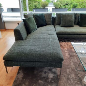 Graues Charles Carré Sofa mit grünen Kissen auf einem braunen Teppich neben einem Couchtisch aus Glas in einem modernen Wohnzimmer mit großen Fenstern - ein echtes Designersofa von B&B Italia.