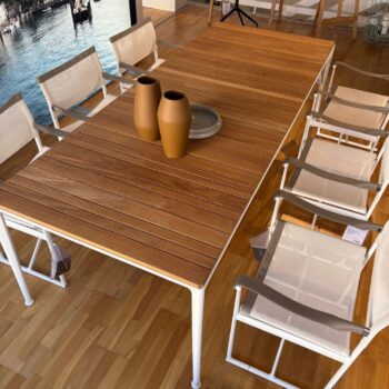 Ein Esstisch aus Teakholz mit weißen Metallbeinen, umgeben von sechs weißen Netzstühlen nach dem Vorbild des Mirto Outdoortischs von B&B Italia. Zwei braune Vasen und ein graues Tablett stehen auf dem Holzboden im Innenbereich.