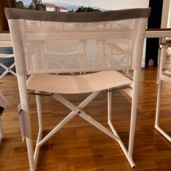 Ein weißer Klappsessel mit Metallrahmen und beigem Netzsitz und Rückenlehne aus der Mirto Outdoor-Kollektion von B&B Italia steht auf einem Holzboden in der Nähe eines Tisches in einem sonnendurchfluteten Raum.