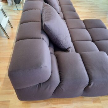 Ein modernes Tufty-Time-Sofa von B&B Italia, gepolstert in Dunkelgrau mit plüschigen, getufteten Kissen, steht auf einem hellen Holzboden neben einem eleganten Glasbeistelltisch.