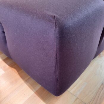 Nahaufnahme der Ecke eines dunkelvioletten Stoff-Ottomanen, der an das ikonische Tufty-Time Sofa von B&B Italia erinnert, auf einem hellen Holzboden.