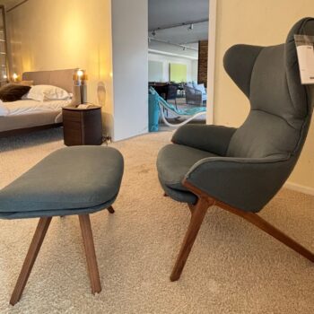 Ein moderner blauer Cassina Sessel mit Holzbeinen und ein passender Cassina Hocker stehen auf einem Teppichboden in einem Schlafzimmer mit einem sichtbaren Bett und Nachttisch im Hintergrund.
