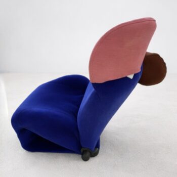 Ein moderner Wink-Stuhl von Cassina mit blauer Sitzfläche, rosafarbener Rückenlehne und brauner, runder Kopfstütze steht auf einem hellen Boden vor einem unifarbenen Hintergrund - ein echter Klassiker-Sessel im zeitgenössischen Design.