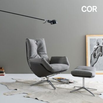 Ein moderner COR Sessel mit passender Ottomane steht auf einem Teppich, neben einer Stehlampe und einem gerahmten Kunstwerk, vor einer schlichten grauen Wand. Das Wort COR erscheint in der oberen rechten Ecke.