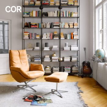 Eine moderne Leseecke mit einem hellbraunen COR Sessel aus Leder und einer Ottomane, einem großen Bücherregal mit Büchern, einer Gitarre, einem Globus und Vinylplatten auf einem hellgrauen Teppich.