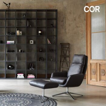 Modernes Wohnzimmer mit einem eleganten COR Sessel und einer Ottomane, einem großen schwarzen Regal mit Dekorationsartikeln und einer Holztür mit Glasfüllung.