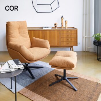Ein modernes Wohnzimmer mit einem hellbraunen Cordia-Drehsessel und einer passenden Ottomane auf einem gemusterten Teppich, einem hölzernen Sideboard und dekorativen Gegenständen im Hintergrund.