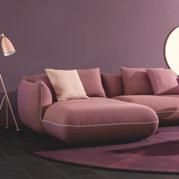 Ein modernes mauvefarbenes JALIS-Sofa mit beigen und mauvefarbenen Kissen steht auf einem dunkelvioletten Teppich, neben einer Stehlampe und einer runden Leuchte an einer mauvefarbenen Wand.