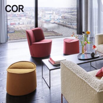 Eine moderne Lounge mit roten und gelben Hockern, Büchern, Blumen und weißen, strukturierten COR-Nenou-Sesseln, die auf ein großes Fenster ausgerichtet sind.