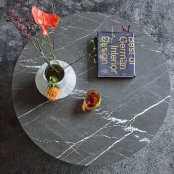 Runder Sting-Marmor-Beistelltisch mit einer weißen Blumenvase, einem Glas Getränk und dem Buch Best of German Interior Design von COR auf einem grauen Teppichboden.