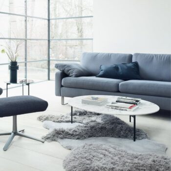 Ein modernes Wohnzimmer mit einem grauen COR-Sofa, einem passenden Sessel, einem runden Marmor-Couchtisch mit Büchern und einem grauen Zottelteppich auf hellem Holzboden neben großen Fenstern und einem schlichten Beistelltisch.