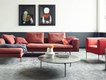Modernes Wohnzimmer mit rotem COR Sofa und Sessel, rundem Beistelltisch, grauem Teppich und zwei gerahmten Porträts an einer weiß-grauen Wand.