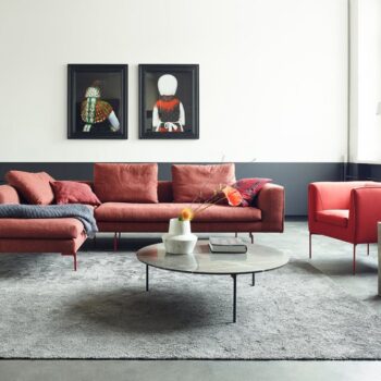 Modernes Wohnzimmer mit rotem COR Sofa und Sessel, rundem Beistelltisch, grauem Teppich und zwei gerahmten Porträts an einer weiß-grauen Wand.