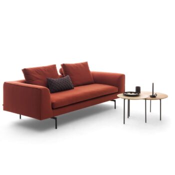 Ein modernes rotes COR-Sofa mit zwei Sitzkissen und drei Wurfkissen steht neben zwei runden, verschachtelten Beistelltischen mit Tablett, Tasse und Kerze auf weißem Hintergrund.