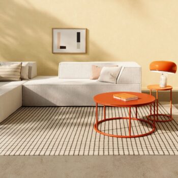 Ein modernes Wohnzimmer mit einem COR-Sofa, einem karierten Teppich, zwei runden orangefarbenen Tischen, einer orangefarbenen Lampe und abstrakter Wandkunst vor einer hellgelben Wand.