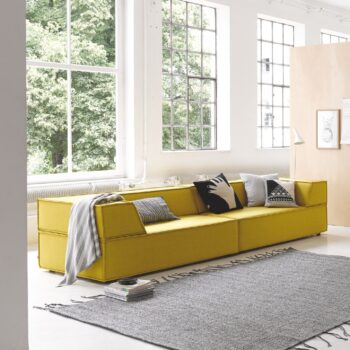 Ein modernes Wohnzimmer mit einem COR-Sofa-Trio in Gelb, verschiedenen Kissen, einem grauen Teppich, großen Fenstern, einer Stehlampe und Kunstwerken an hellen Wänden.
