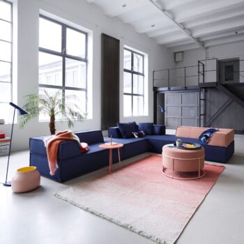 Modernes Wohnzimmer mit blauem COR Sofa Trio, rosafarbenen Sesseln, rundem Couchtisch, Stehlampen, Topfpalme, großen Fenstern und einem Loft mit Treppe, das die Eleganz der COR-Möbel unterstreicht.