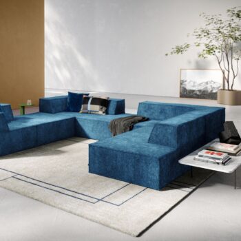 Ein modernes Wohnzimmer mit einem großen blauen COR-Sofa, einem hellen geometrischen Teppich, einem Marmor-Couchtisch mit Büchern und einer Topfpflanze neben einem gerahmten Bergfoto an der Wand.