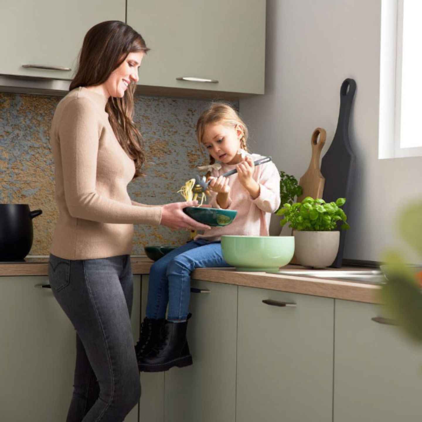 Ein Erwachsener reicht einem Kind, das auf einem Küchentisch sitzt, eine Schüssel mit Nudeln. Das Kind benutzt eine Zange, und auf dem Tresen sind frische Kräuter zu sehen. Ein Erwachsener reicht einem Kind, das auf einem Küchentisch sitzt, eine Schüssel mit Nudeln. Das Kind benutzt eine Zange, und auf dem Tresen sind frische Kräuter zu sehen.